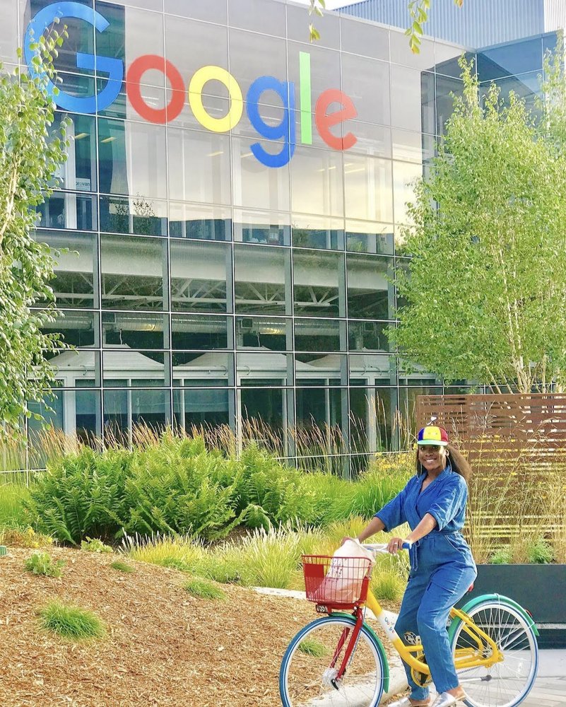 Ada, wearing a Noogler hat and sitting on a Google bike, poses in front of the Google logo at our Mountain View HQ