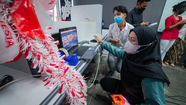 An Indonesian woman with a face mask is having a video conference call on her laptop. In the background, a fellow Indonesian man with a face mask is observing the call.