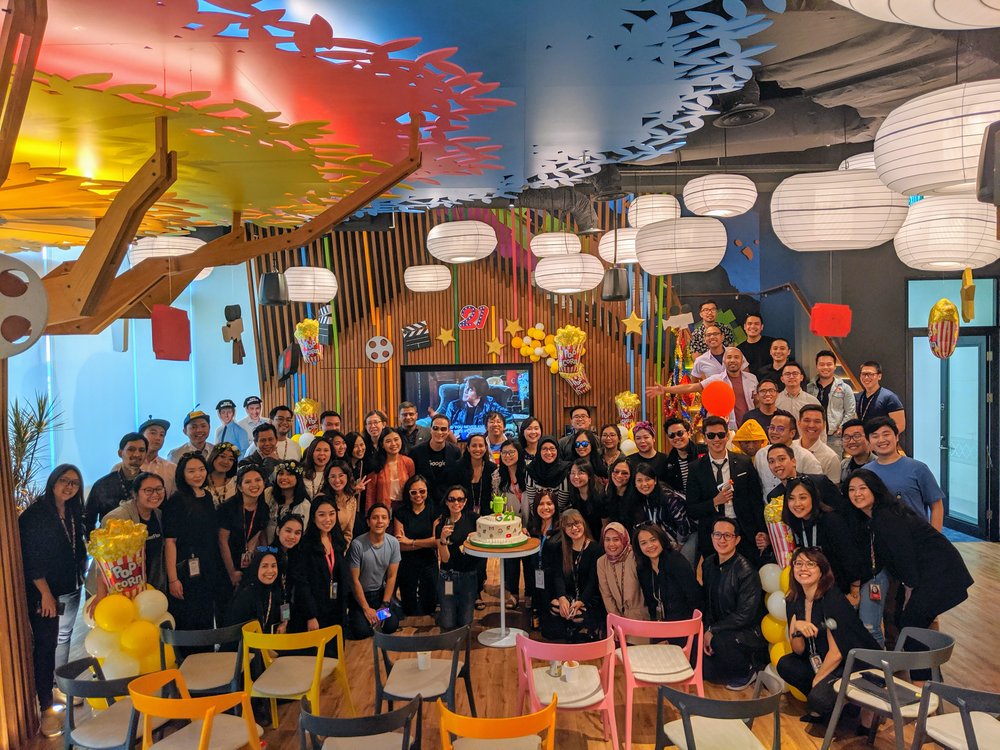 A group photo of male and female Google employees at the office cafe smiling and dressed up in party gear, celebrating Google’s global 21st anniversary.