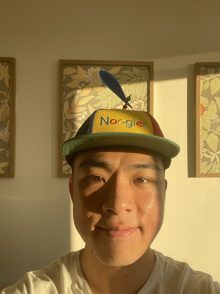 Ken smiling in his Noogler hat. Sunlight is pouring in from a nearby window.