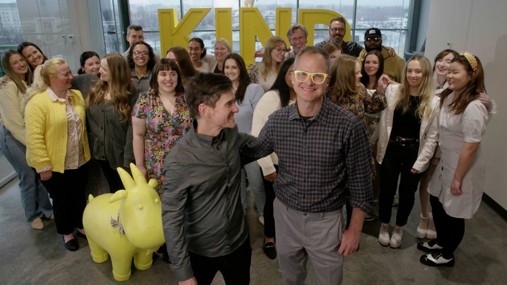 Two men stand in front of a group of smiling people, the word “KIND” in big yellow letters on the window behind them all, and a yellow model of a goat.