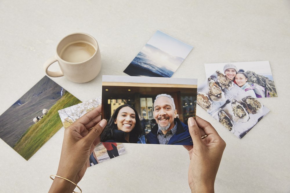 Two hands holding a photo of a smiling couple, other photos and a cup of coffee in the background.