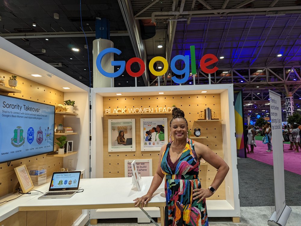 A woman in a multicolored dress stands in a booth under a Google sign, with her hand resting on a table.