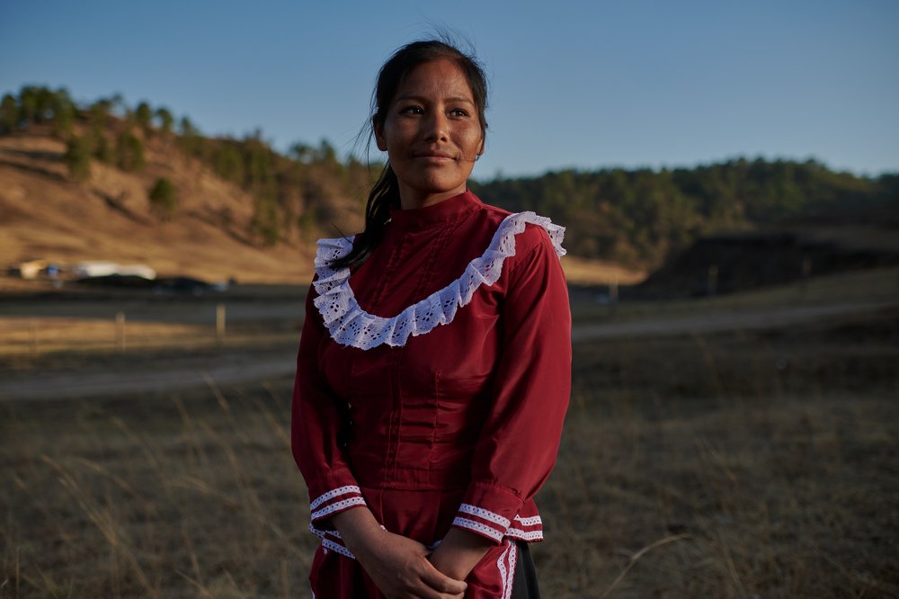 A Mexican woman wearing a red dress with a white ruffle stands in front of hills, looking slightly away from the camera.