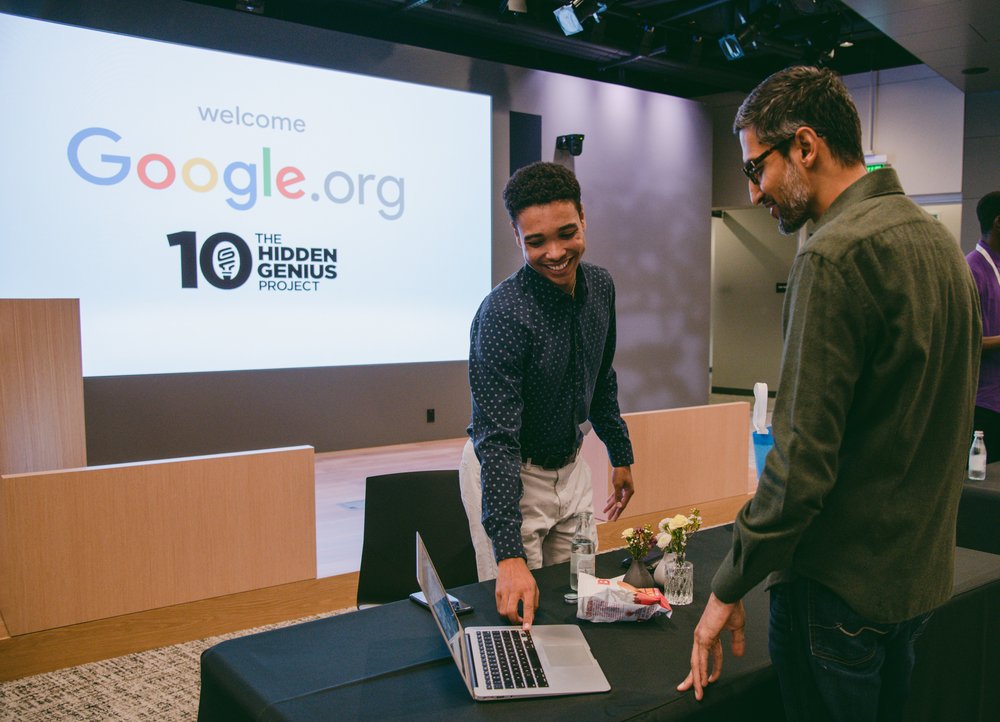 Student Ian stands behind a desk operating a laptop to demonstrate his platform for Sundar, who is standing to the right of the desk. Both are smiling. A screen in the background shows the Google.org and The Hidden Genius Project logos.